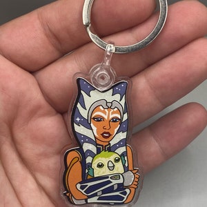 May include: A clear acrylic keychain featuring a cartoon illustration of Ahsoka Tano from Star Wars, holding a small green and yellow Loth-cat. The background is a purple and blue galaxy pattern.
