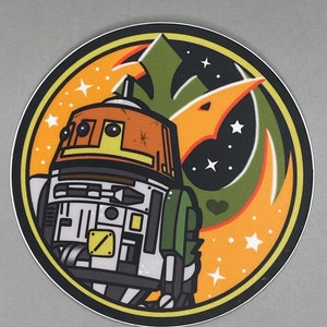 May include: A round sticker with an orange, green, and black design. The sticker features a cartoon image of a droid with a yellow and orange head, a white body, and a green arm. The droid is surrounded by a stylized orange and green symbol with white stars.