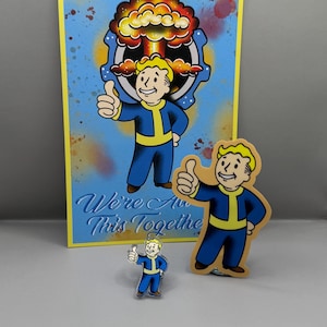 May include: A set of three Fallout Vault Boy themed items. A large print with a blue background, a yellow Vault Boy with a thumbs up, and a mushroom cloud. A smaller sticker with a yellow Vault Boy with a thumbs up. A small pin with a yellow Vault Boy with a thumbs up. The print has the text "We're All This Together".