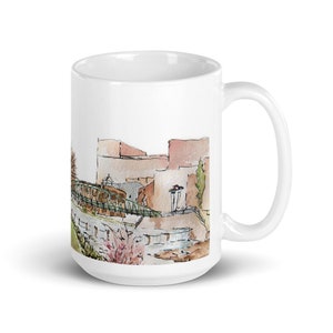 May include: White ceramic mug with a watercolor illustration of a bridge and buildings. The illustration is in shades of brown, green, and pink.