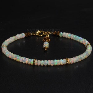 Op de afbeelding: Een opaalarmband met een gouden sluiting en accenten. De armband heeft kleine, iriserende opaal kralen, afgewisseld met goudkleurige kralen. Een kleine bedel hangt aan de sluiting. De armband wordt getoond tegen een donkere achtergrond.