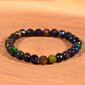 Peut inclure: Un bracelet de perles d'opale facettées de couleurs sombres. Les opales présentent des reflets verts, rouges et bleus, avec une finition polie. Le bracelet est présenté sur une surface brun clair. Les perles mesurent environ 0,8 cm de diamètre.