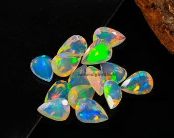 May include: A collection of pear-shaped opals with iridescent colors, including blue, green, and yellow. The gemstones are scattered on a black surface, with a wooden element in the background. The text "naturalgemshub" is visible.