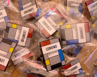 May include: A close-up of several colorful enamel pins with a geometric design. The pins feature the word "LEMOND" and the phrase "La Vie Claire" in white text. The pins are packaged in clear plastic bags.