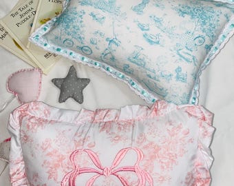 Può includere: Due cuscini decorativi, uno con un motivo floreale blu e bianco e l'altro con un motivo floreale rosa. Il cuscino rosa ha un fiocco rosa e il nome "Leah" ricamato.