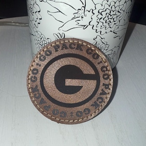 May include: A brown leather coaster with a stitched edge and a black circle with the text "Go Pack Go" and a black letter "G" in the center.