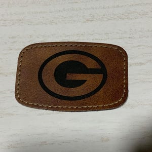 May include: A brown leather patch with a stitched border and a black letter "G" inside a circle.