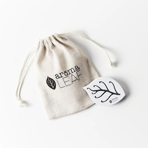 Könnte beinhalten: Eine weiße Kordeltasche mit dem aufgedruckten Text "aroma LEAF". Neben der Tasche liegt ein weißes Keramikblatt mit einem schwarzen Astdesign und dem aufgedruckten Text "aroma LEAF".