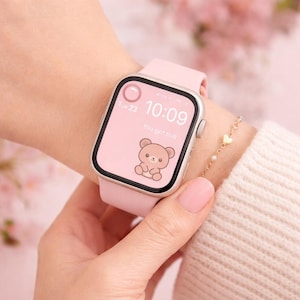 Pode incluir: Um smartwatch rosa com uma pulseira a condizer mostra as horas 10:09 e um gráfico de urso fofo. O relógio é usado num pulso, com uma pulseira dourada com berloques de coração. O ecrã também mostra o texto "You got this".