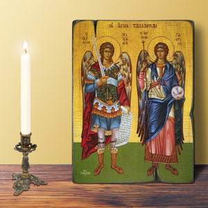 May include: An icon depicting two figures with halos, holding a sword and a staff, with text at the top. The figures are dressed in robes with gold accents. A lit white candle sits in a decorative brass candlestick.