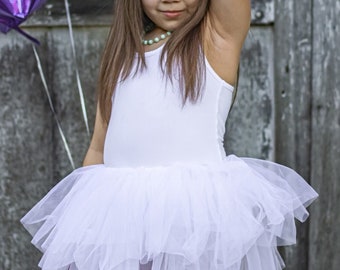 May include: A young person wearing a white leotard and a layered white tulle skirt, holding pink balloons. The background includes purple and white balloons, and a weathered wooden wall.