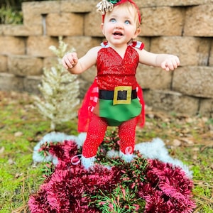 May include: A baby girl wearing a red and green elf costume with a sparkly red top, green skirt, and white tights. She is standing on a pile of red and green tinsel.