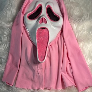 May include: A white Scream mask with black eye holes and a pink glitter-filled mouth, set against a pink hooded robe. The mask is a classic Halloween costume element, with a simple, yet eerie design.