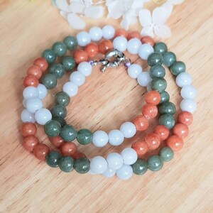 May include: A multi-strand necklace made of jade beads in three colors: white, green, and orange. The beads are round and smooth, and the necklace is strung on a silver chain.