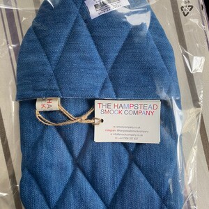 Pode incluir: Uma capa para garrafa de &aacute;gua quente de ganga azul com um padr&atilde;o acolchoado de losangos. A capa est&aacute; embalada num saco de pl&aacute;stico transparente com um r&oacute;tulo que diz "The Hampstead Smock Company" e inclui informa&ccedil;&otilde;es de contacto.