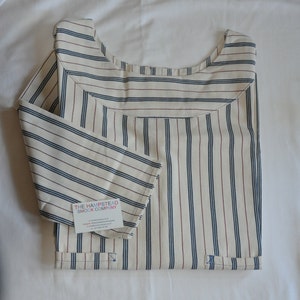 May include: A white and blue striped smock with red stripes. The smock has a round neckline and is folded in half. A small white label with the text "The Hampstead Smock Company" is attached to the smock.
