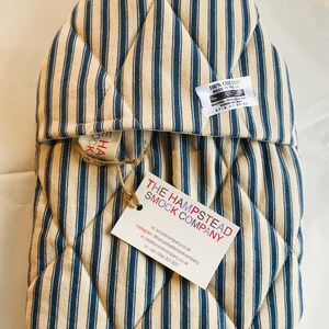 Pode incluir: Um pegador de panela acolchoado em azul e branco listrado com uma etiqueta que diz "The Hampstead Smock Company" e inclui informa&ccedil;&otilde;es de contato. A etiqueta est&aacute; presa com um barbante.