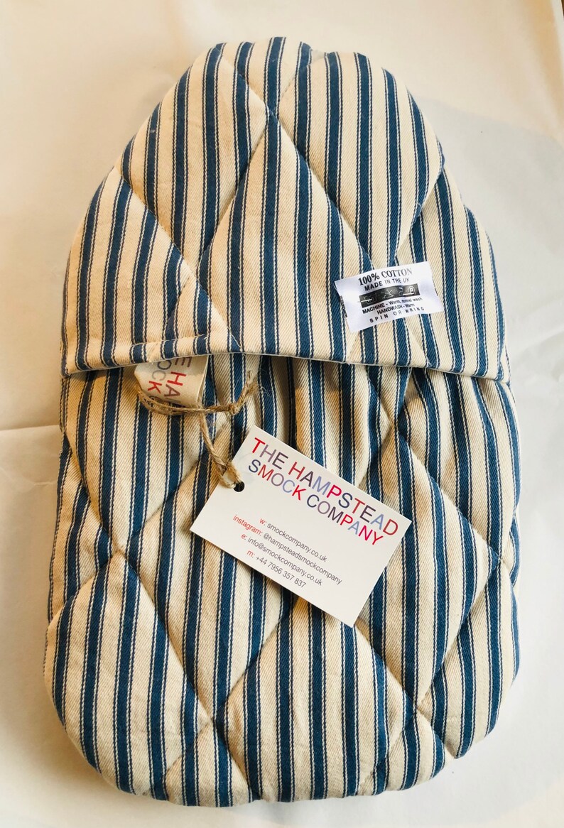 Pode incluir: Um pegador de panela acolchoado em azul e branco listrado com uma etiqueta que diz "The Hampstead Smock Company" e inclui informa&ccedil;&otilde;es de contato. A etiqueta est&aacute; presa com um barbante.