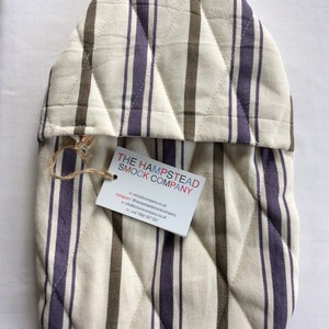 Pode incluir: Uma luva de forno acolchoada com listras marrons e roxas. A luva tem uma etiqueta anexada com o texto "THE HAMPSTEAD SMOCK COMPANY" e informa&ccedil;&otilde;es de contato.