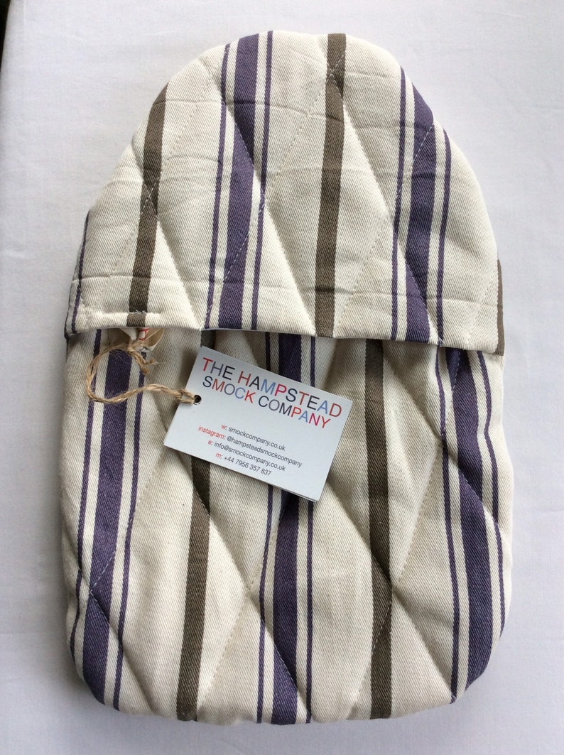 Pode incluir: Uma luva de forno acolchoada com listras marrons e roxas. A luva tem uma etiqueta anexada com o texto "THE HAMPSTEAD SMOCK COMPANY" e informa&ccedil;&otilde;es de contato.