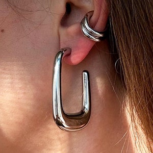 May include: Silver-toned, rectangular hoop earrings and a matching ear cuff. The earrings have a polished, reflective surface and a modern, minimalist design. The ear cuff features a layered design. The jewelry is worn on an ear.