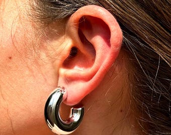 May include: Silver hoop earrings with a thick, rounded design. The earrings are worn in a person's ear.