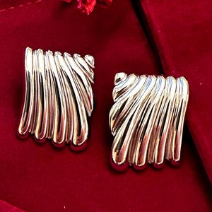 May include: A pair of silver tone earrings with a wavy, fan-like design. The earrings are on a red velvet background.