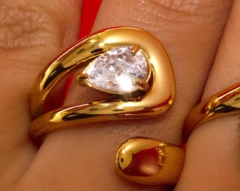 Könnte beinhalten: Zwei goldfarbene Ringe mit großen, klaren Edelsteinen. Ein Ring hat einen tropfenförmigen Stein, der andere einen quadratischen Stein. Die Ringe haben ein modernes, offenes Design mit glatter, polierter Oberfläche.