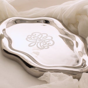 May include: A silver compact mirror with a decorative design on the lid. The mirror is slightly curved and has a smooth, reflective surface.