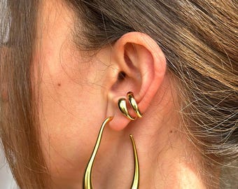 May include: A person wearing a pair of gold hoop earrings. The earrings are large and have a unique teardrop shape.