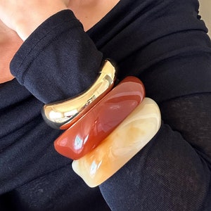 May include: A collection of four bracelets in varying colors and materials. The top bracelet is gold-toned, followed by a reddish-brown, and a creamy white. The bracelets are stacked on a person's arm, against a black long-sleeve top.