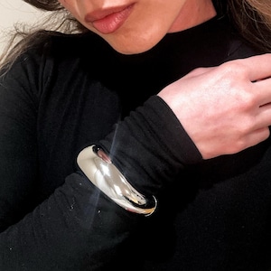 Könnte beinhalten: Ein silbernes, glattes, gebogenes Armreif-Armband. Das Armband wird am Handgelenk einer Person getragen, die ein schwarzes Langarmshirt trägt.