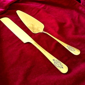 May include: A set of two gold-toned cake servers with ornate designs. The servers are on a red velvet background.