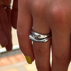 Op de afbeelding: Een zilveren ring met een uniek, golvend ontwerp. De ring wordt gedragen op de middelvinger van een hand met gele nagellak.
