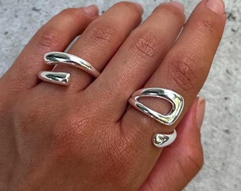 May include: Two silver rings on a hand. One ring is a simple band with a twist, the other is a more abstract design with a cutout.