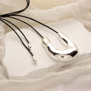 Op de afbeelding: Een zilveren abstracte hangerketting met een unieke, organische vorm. De ketting is aan een zwart leren koord.