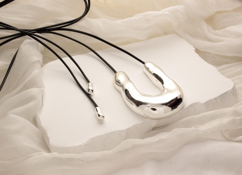 Op de afbeelding: Een zilveren abstracte hangerketting met een unieke, organische vorm. De ketting is aan een zwart leren koord.
