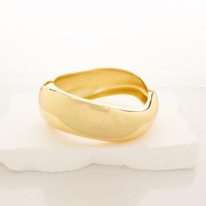 May include: A gold-colored bangle bracelet with a smooth, rounded, and slightly curved design.