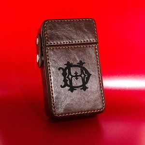 May include: A brown leather cigarette case with a black monogram of the letters "HD" in a decorative font.