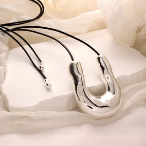 Op de afbeelding: Een zilveren ketting met een grote, abstracte, U-vormige hanger. De hanger heeft een glad, glanzend oppervlak en is bevestigd aan een zwart leren koord.