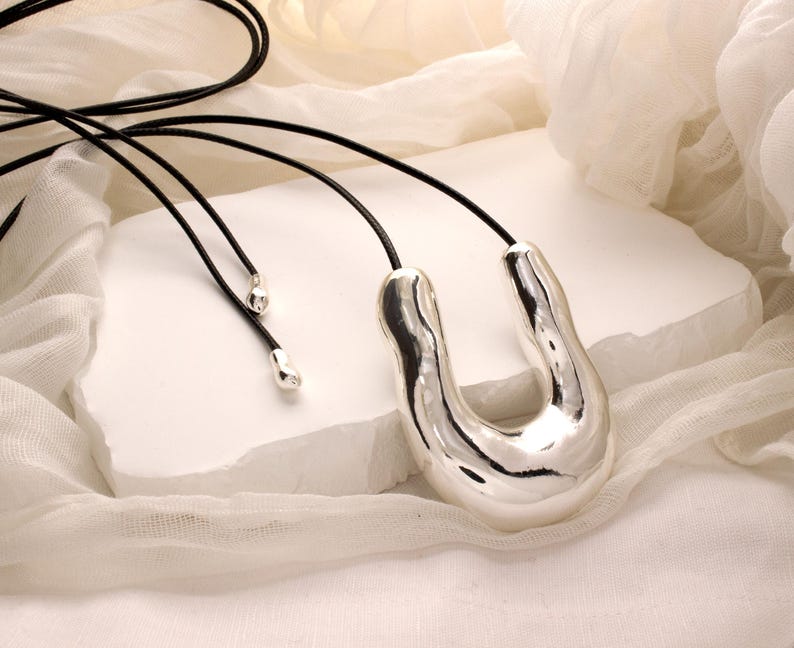 Op de afbeelding: Een zilveren ketting met een grote, abstracte, U-vormige hanger. De hanger heeft een glad, glanzend oppervlak en is bevestigd aan een zwart leren koord.