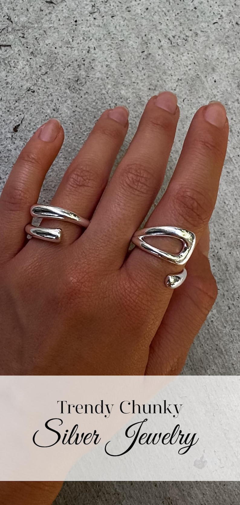 May include: Two silver rings on a hand. The ring on the index finger is a chunky, open ring with a triangular shape. The ring on the middle finger is a thick, twisted band.