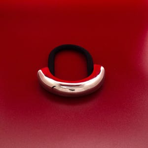 Op de afbeelding: Een zilveren metalen haarband met een glad, afgerond oppervlak. De haarband ligt op een rode achtergrond.