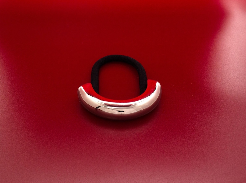 Op de afbeelding: Een zilveren metalen haarband met een glad, afgerond oppervlak. De haarband ligt op een rode achtergrond.