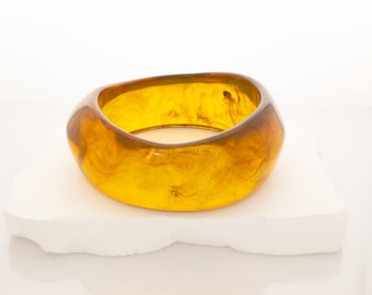 May include: A wide, amber-coloured bracelet with a marbled effect. The oval-shaped bangle is made of a translucent material, displaying golden and brown hues. The bracelet is displayed on a white, textured surface.