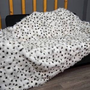 May include: A white faux fur throw blanket with black spots is draped over a black sofa. The blanket has a soft, plush texture and a dalmatian-like pattern. The throw adds a touch of luxury and warmth to the living room.