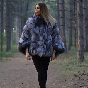 May include: A woman wearing a silver fox fur coat with black trousers and black heels. She is standing in a woodland setting.
