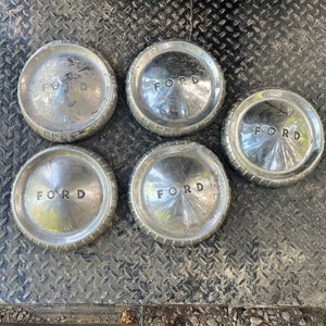 Puede incluir: Cinco tapacubos plateados con la palabra "FORD" en relieve. Los tapacubos están dispuestos en círculo sobre una superficie metálica negra.