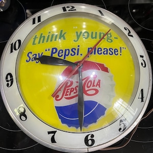 May include: A vintage Pepsi-Cola clock with a yellow background and a white border. The clock face features the Pepsi logo and the phrase "think young - Say 'Pepsi, please!'". The clock hands are black and red.