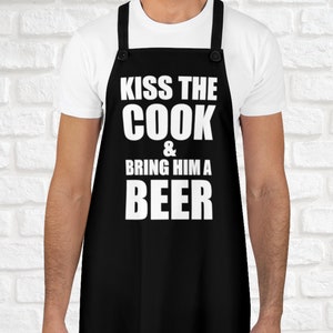 Könnte beinhalten: Schwarze Schürze mit weißem Text, der "Kiss the Cook & Bring Him a Beer" lautet.